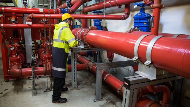 Engineer in high-visibility jacket inspecting red fire-protection pipework in a building plant room