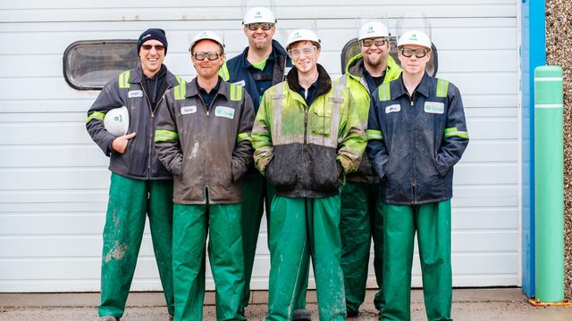Team of six uniformed maintenance workers in hard hats standing together in front of a service bay