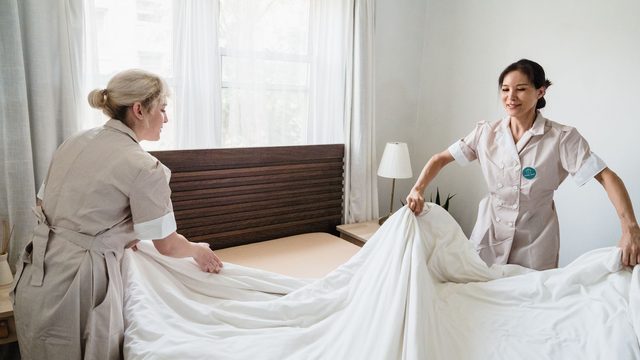 Two hotel housekeepers in uniform making up a bed with fresh linen in a guest room