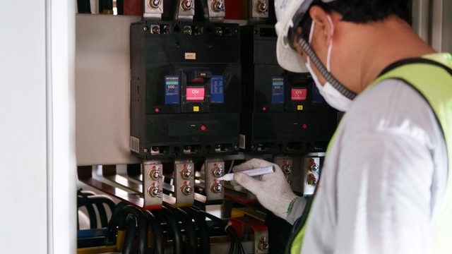 Technician in white helmet recording readings on a clipboard while inspecting an electrical switchboard